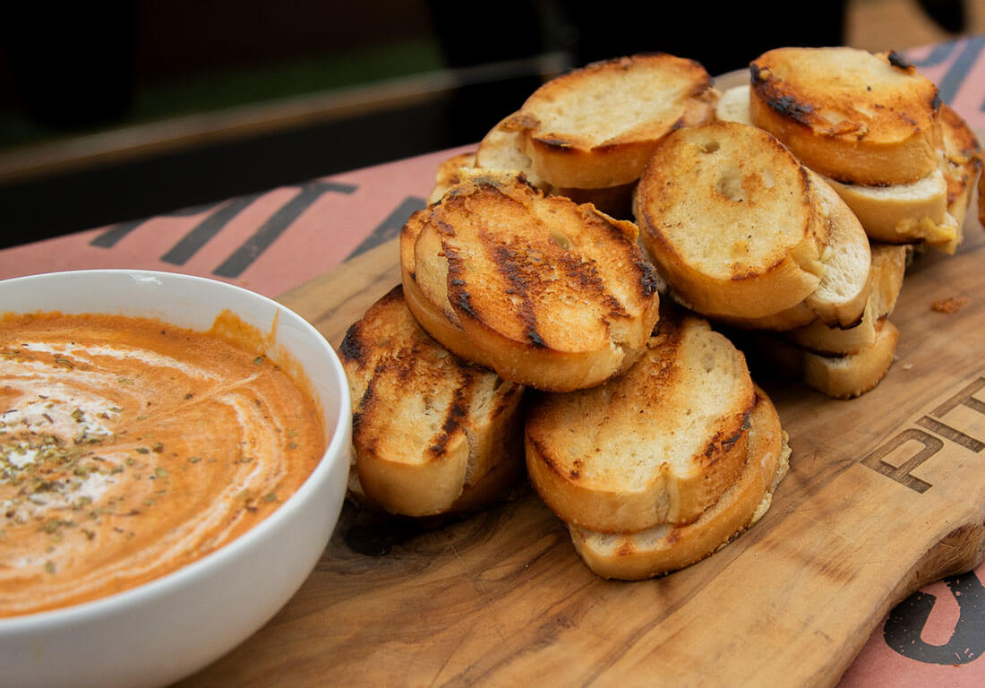 crispy grilled cheese cooked on flat top gas griddle paired with smoked and roasted tomato soup cooked on pit boss wood pellet grill and served in white bowl and wooden cutting board